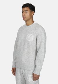 Jasnoszary, dziany sweter z okrągłym dekoltem, ściągaczami na rękawach i logo wyszytym na piersi. Miękka tekstura i luźny krój.