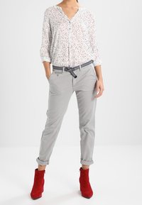 Blusa leggera a pois con scollatura a V e maniche arrotolate, abbinata a pantaloni grigi e stivaletti alla caviglia in suede rossi, accentuata da una cintura a fantasia.