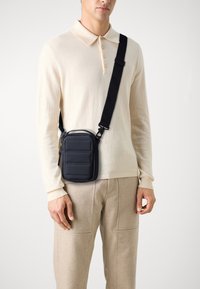 Schwarze gepolsterte Umhängetasche in rechteckiger Form, mit einem Reißverschluss und verstellbarem Riemen, getragen über einem hellcremefarbenen Pullover.
