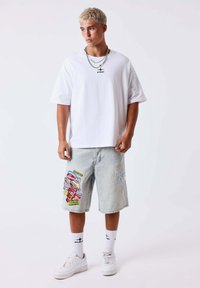 Joven con el pelo rubio platino corto lleva una camiseta blanca extragrande, pantalones cortos vaqueros claros con parches de colores, calcetines blancos y zapatillas blancas.