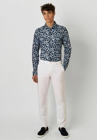 Blau-weißer gemusterter Button-up-Hemd mit langen Ärmeln, kombiniert mit hellen Hosen und schwarzen Slippern, vor einem neutralen Hintergrund stehend.