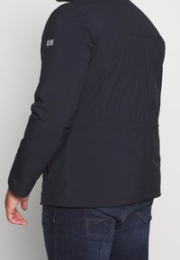 Tmavě modrá, zateplená bunda hladké textury, s logem na levém rukávu a bočními kapsami, navržená pro teplo a pohodlí.