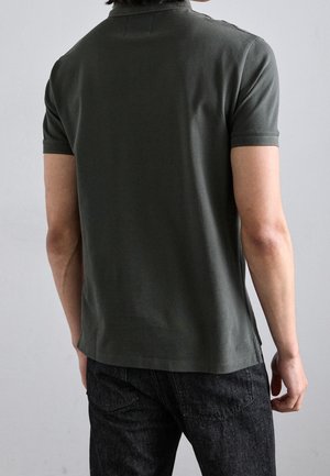 Homme portant un polo vert foncé à manches courtes et un jean noir, vu de dos sur un fond gris clair uni.