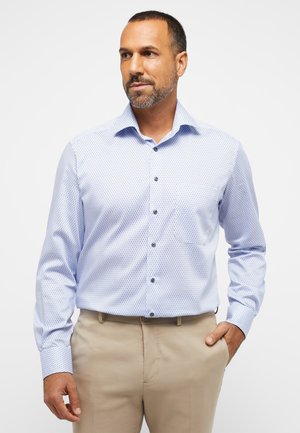 Helle blaue Hemdbluse mit kleinen dunkelblauen Pünktchen, langen Ärmeln und einer Brusttasche, kombiniert mit beigen Hosen.