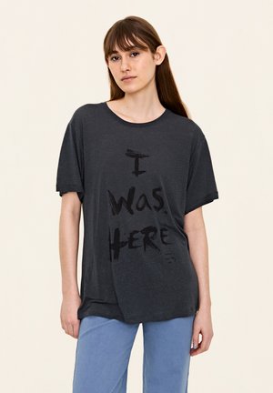 T-shirt en coton gris foncé à manches courtes et col rond. Présente un impression graphique noire avec le texte "J'étais ici" à l'avant.