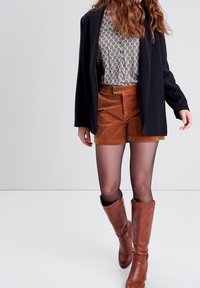 Černý blejzr přes vzorovanou světlou košili, doplněný hnědými kordovým šortkami. Hnědé vysoké kozačky, na nohou jsou viditelné průsvitné černé punčocháče.