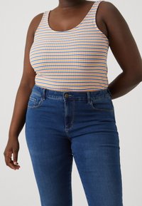 Randig ärmlös topp i orange, blå och gul tyg; matchad med mörkblå tajta jeans med framfickor och knäppning.