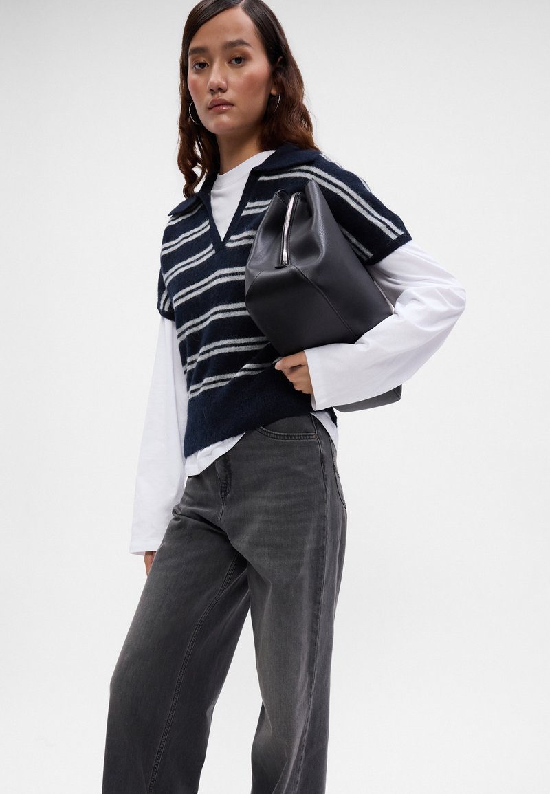 Niebiesko-białe prążkowane kamizelka dzianinowa nałożona na białą koszulę z długim rękawem, dopasowana do szarych spodni dzwonów. Model trzyma czarną torbę na zamek błyskawiczny.