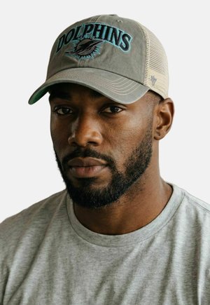 Mann mit Bart, der eine graue Netzrückenkappe mit dem "Dolphins"-Logo und ein hellgraues T-Shirt mit Rundhalsausschnitt trägt, schaut vorwärts vor weißem Hintergrund.