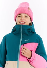 Pestrá zimní bunda s tyrkysovými, růžovými a béžovými panely. Obsahuje vysoký límec, kapsu na zip a nastavitelné manžety. Na hlavě má růžovou pletenou čepici.