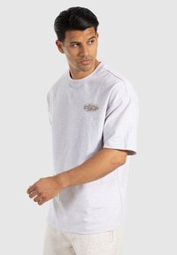 Camiseta de manga corta gris claro, hecha de una mezcla de algodón, con cuello redondo y un pequeño logo bordado en el pecho. Textura suave.