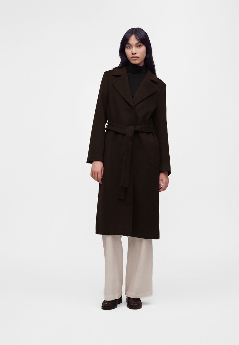 Femme aux longs cheveux foncés portant un long manteau marron foncé ceinturé, un haut noir, un pantalon large crème et des chaussures marron foncé, debout devant un fond uni.