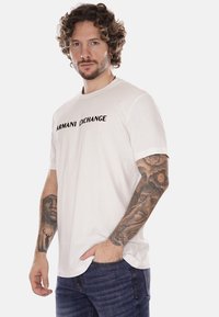 T-shirt bianco in cotone con maniche corte, con il logo nero "ARMANI EXCHANGE" sulla parte anteriore. Indossato con jeans blu.