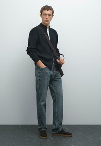 Giovane uomo che indossa un maglione nero con zip, jeans blu e scarpe scure, in piedi con una mano in tasca e mentre tiene la tracolla di una borsa.