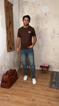 Άνδρας με κοτσίδες, φορώντας καφέ μπλουζάκι "Osaka", μπλε τζιν και λευκά αθλητικά παπούτσια, στέκεται σε ρουστίκ δωμάτιο με ξύλινο πάτωμα και δίπλα του υπάρχει δερμάτινη τσάντα.