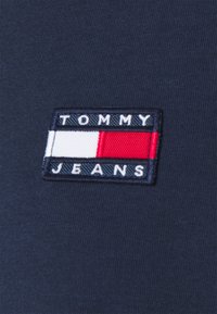Tumma laivansinisestä puuvillakankaasta valmistettu suorakaiteen muotoinen brodeerattu paikka, jossa on valkoinen teksti "TOMMY" punavalkoisen suorakaiteen yläpuolella ja "JEANS" valkoisena alapuolella.