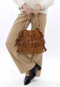 Brun poncho-taske lavet af ruskind med runde sølvnagler og snorelukning, holdt af en person i en cremet sweater og beige bukser.