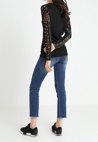 Pull noir avec manches longues à motifs, associé à un jean bleu slim et des baskets noires. La vue de dos met en valeur les motifs des manches et la texture.