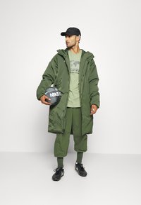 Grüne Parkajacke mit Kapuze, getragen über einem hellgrünen T-Shirt, kombiniert mit grünen Hosen und schwarzen Schuhen; hält einen schwarzen Nike-Fußball.
