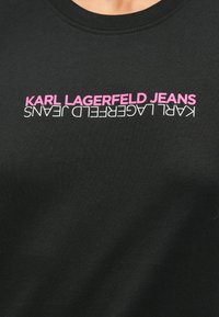 Melns kokvilnas sporta krekls ar baltu un rozā "KARL LAGERFELD JEANS" teksta dizainu, horizontālā alinejā, ar apaļu kakla izgriezumu un mīkstu tekstūru.