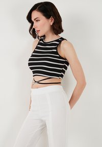 Čierny a biely pruhovaný crop top s priliehavým strihom, s bezrukávovým lemom a krížovými popruhmi v oblasti pása, spárovaný s bielymi nohavicami.