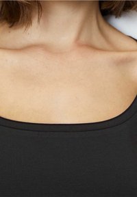 Kvinnas övre bröst och nacke med kort brunt hår, iförd en svart off-shoulder topp.