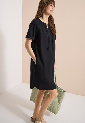 Frau in einem schwarzen kurzärmeligen Kleid mit Taschen, die eine große gesteppte grüne Tragetasche hält, lächelnd und vor einem beigen Hintergrund steht.