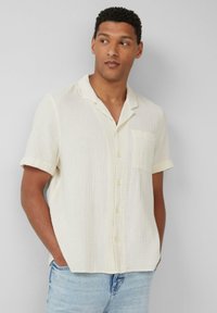 Korte mouwen, lichtbeige shirt van gestructureerde stof, met een knoopsluiting aan de voorkant, kraag en zak. Gecombineerd met lichtblauwe jeans.