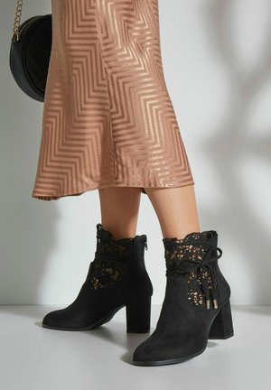 Stivaletti in suede neri con un design in pizzo sulla parte superiore, punta chiusa, zip laterale e tacco spesso, abbinati a un vestito con motivo.