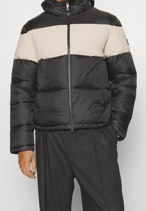 Homme portant une doudoune bicolore noire et beige avec fermeture éclair frontale et poches zippées, portée avec un pantalon gris foncé.