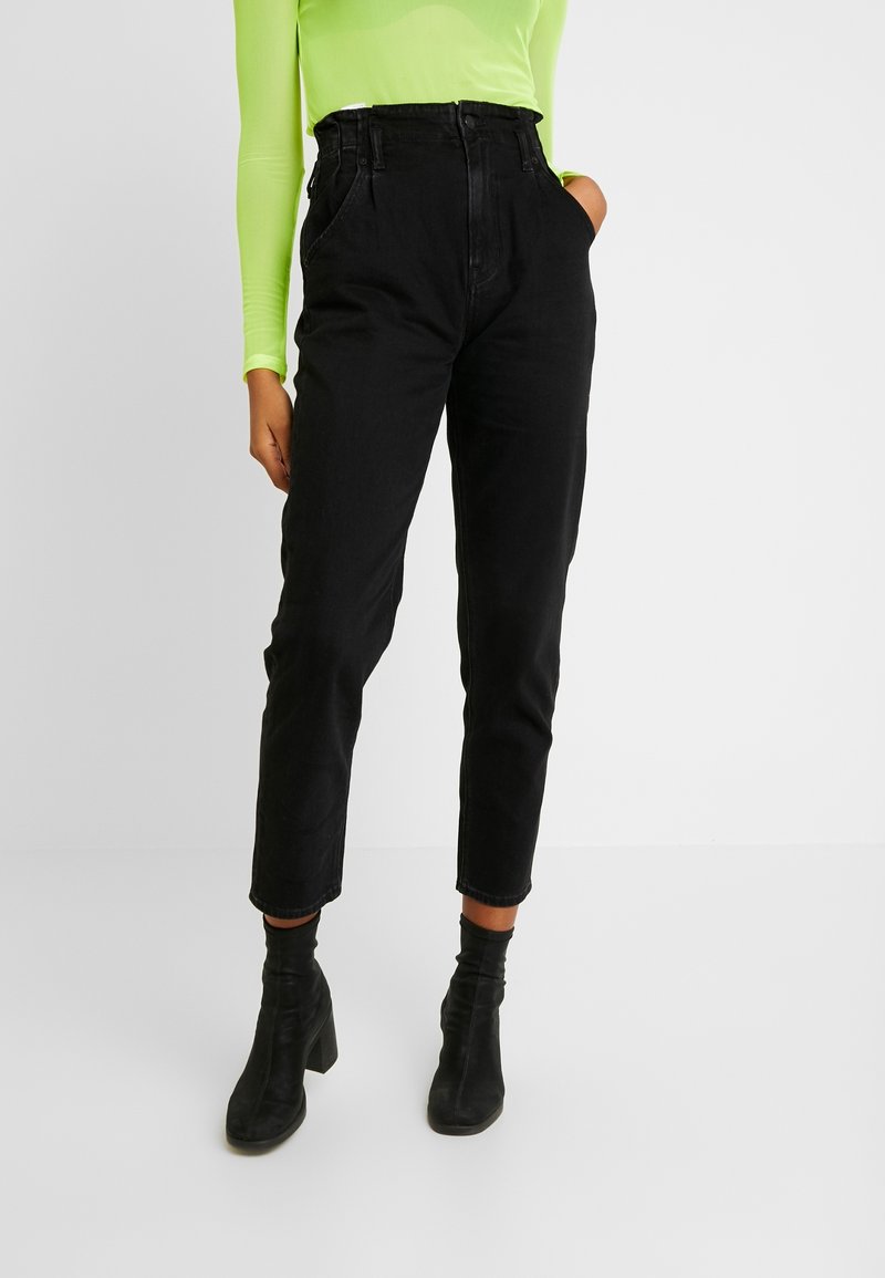 Hoge zwarte denim jeans met een taps toelopende pijp, voorzien van voorkeurszakken en riemlussen. Gedeeld met zwarte hoge enkel laarzen.