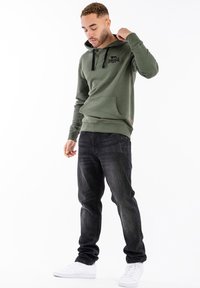 Sudadera verde oliva con cordones negros, bolsillo frontal y logo. Combinada con jeans oscuros y zapatillas blancas, creando un look casual.