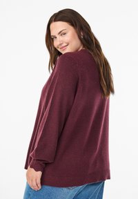 Bordeaux gestrickter Pullover mit langen Ärmeln und gerippten Bündchen. Der Stoff wirkt weich mit einer leicht strukturierten Oberfläche. Lässige Passform, getragen mit blauen Jeans.