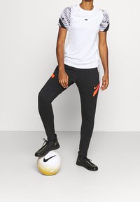 Witte sportshirt met grijze patroonmouwen, gecombineerd met zwarte broek met een oranje logo. Model staat op een witte en gouden voetbal.