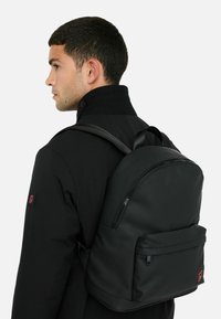 Sort rygsæk med afrundet design, glat tekstur, frontlomme med lynlås og justerbare remme, båret på ryggen over en sort jakke.