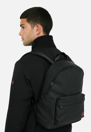 Čierny batoh s zaobleným dizajnom, hladkou textúrou, prednou zipsovou vreckom a nastaviteľnými popruhmi, nosený na chrbte cez čiernu bundu.