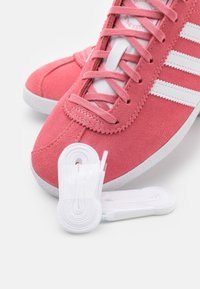 Pink suede sneakers feature white leather three stripes and matching laces. A textured upper complements the white rubber sole.