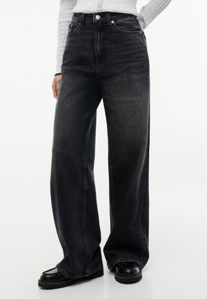 Schwarze, hoch taillierte Jeans mit weitem Bein, getragen mit schwarzen Schuhen und einem hellgrauen gerippten Pullover, von Taille bis Füße gezeigt.