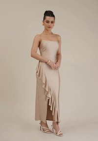 Tettsittende, beige satin-kjole med spaghettistropper, sideryggdetalj og splitt. Kombinert med hvite åpen-tå hæler. Glatt tekstur.