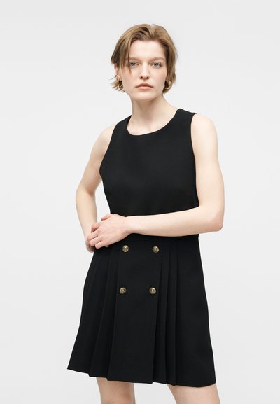 Γυναίκα με κοντά μαλλιά που φοράει μαύρο φόρεμα χωρίς μανίκια με πλισέ φούστα και χρυσά κουμπιά, στέκεται με τα χέρια δεμένα στη μέση.
