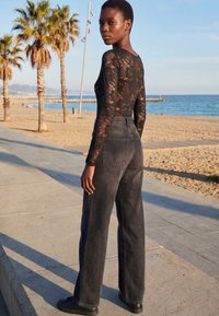 Jeune femme en haut en dentelle noire et jean noir délavé à jambes larges, debout sur une promenade côtière avec des palmiers et l’océan en arrière-plan.