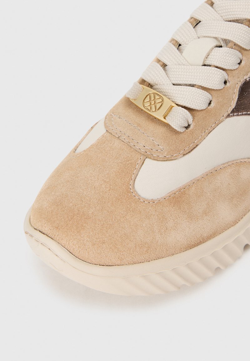 Sneaker in suede tan con accenti crema e marrone, superficie testurizzata, punta rotonda e dettaglio in metallo dorato sui lacci. Suola rigata.