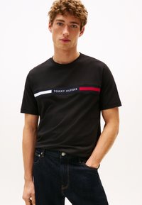 Mladi moški z kodrastimi lasmi nosi  črno majico Tommy Hilfiger z rdečimi, belimi in modrimi črtami ter temno moder kavbojke, z eno roko v žepu.