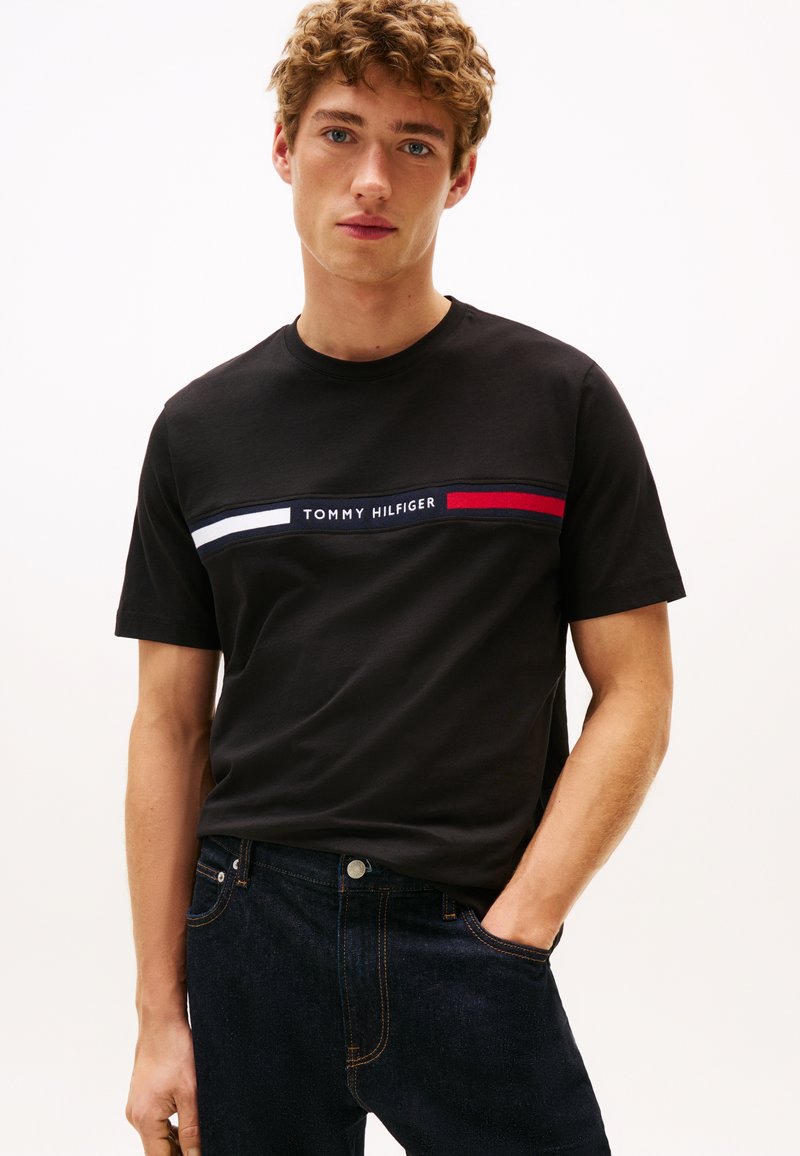 Mladi moški z kodrastimi lasmi nosi črno majico Tommy Hilfiger z rdečimi, belimi in modrimi črtami ter temno moder kavbojke, z eno roko v žepu.