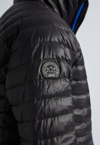 Zwarte pufferjack met een doorgestikt ontwerp, glanzende textuur, blauwe ritsaccenten en een ronde logo patch op de linkermouw.