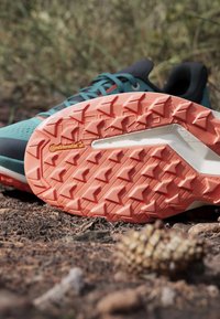 Laufschuh mit orangefarbener Continental-Gummisohle, der auf Waldboden mit Kiefernzapfen und trockenen Blättern liegt.