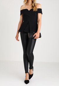 Haut péplum noir à épaules dénudées, leggings noirs brillants ajustés, et escarpins noirs à bout pointu, assortis à une pochette scintillante.