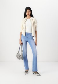 Giacca beige chiaro con zip sopra una maglietta bianca, abbinata a jeans chiari a campana. Tacchi metallici argento e una piccola borsa a spalla argentata.