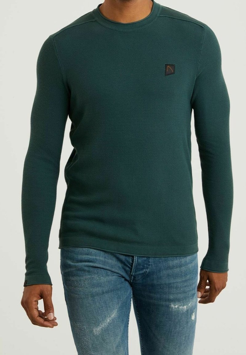 Homme portant un pull texturé vert foncé à manches longues et un jean bleu usé, debout devant un fond blanc.