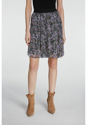 Vrouw met een zwarte knielange rok met een paars bloemenpatroon en beige suède enkellaarsjes, staand op een grijze vloer.